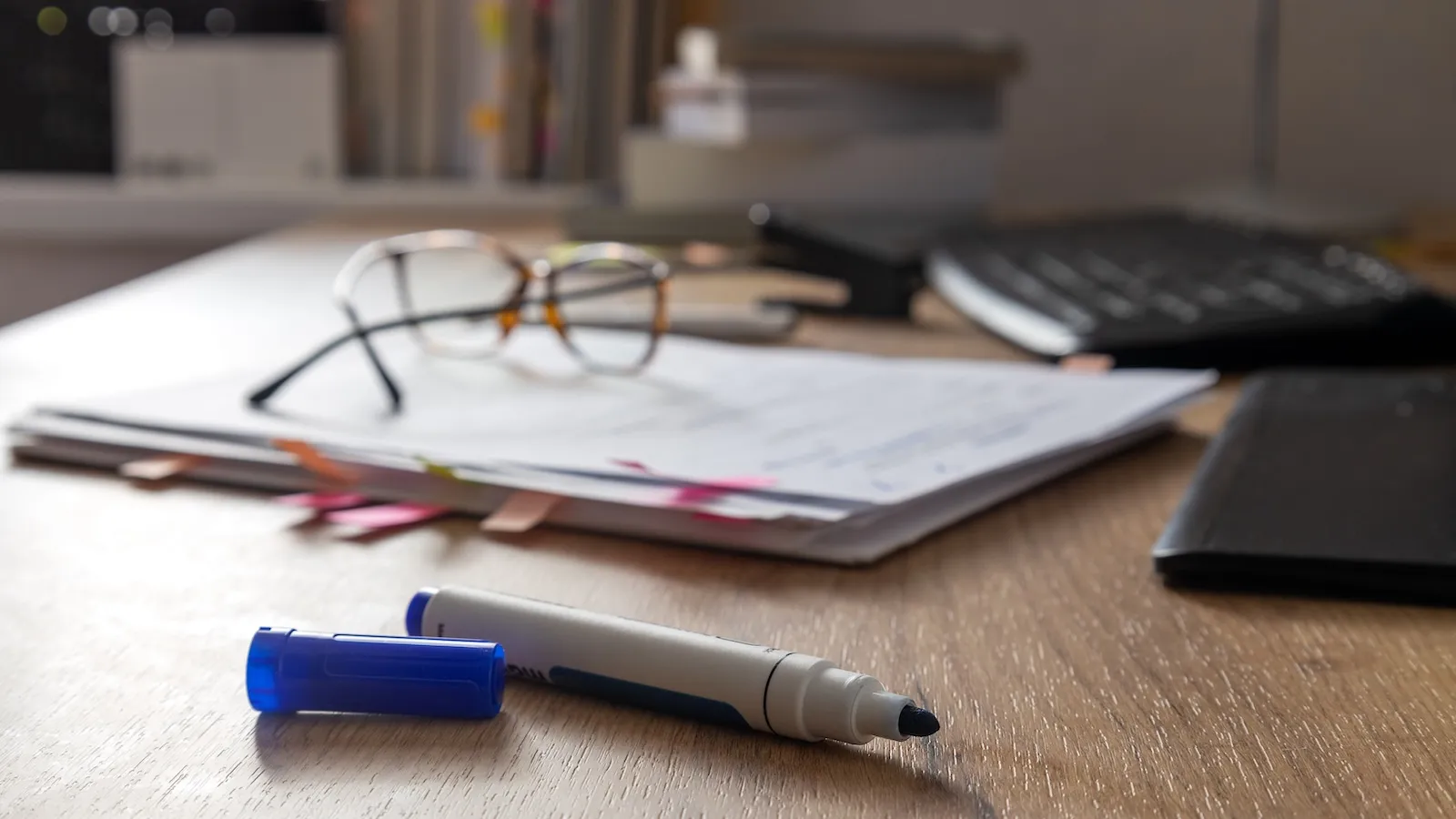 A blue marker, uncapped, sits on a wooden desk, with a pile of papers, a pair of glasses, a keyboard, and a stack of books in the background