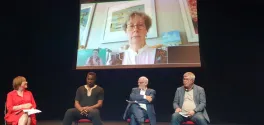 Four presenters sit onstage with one presenter projected on a screen behind them